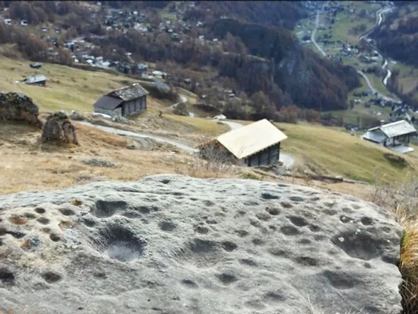 Photographie d’une roche dominant une vallée en montagne avec de multiple cupules, à proximité d’un village.